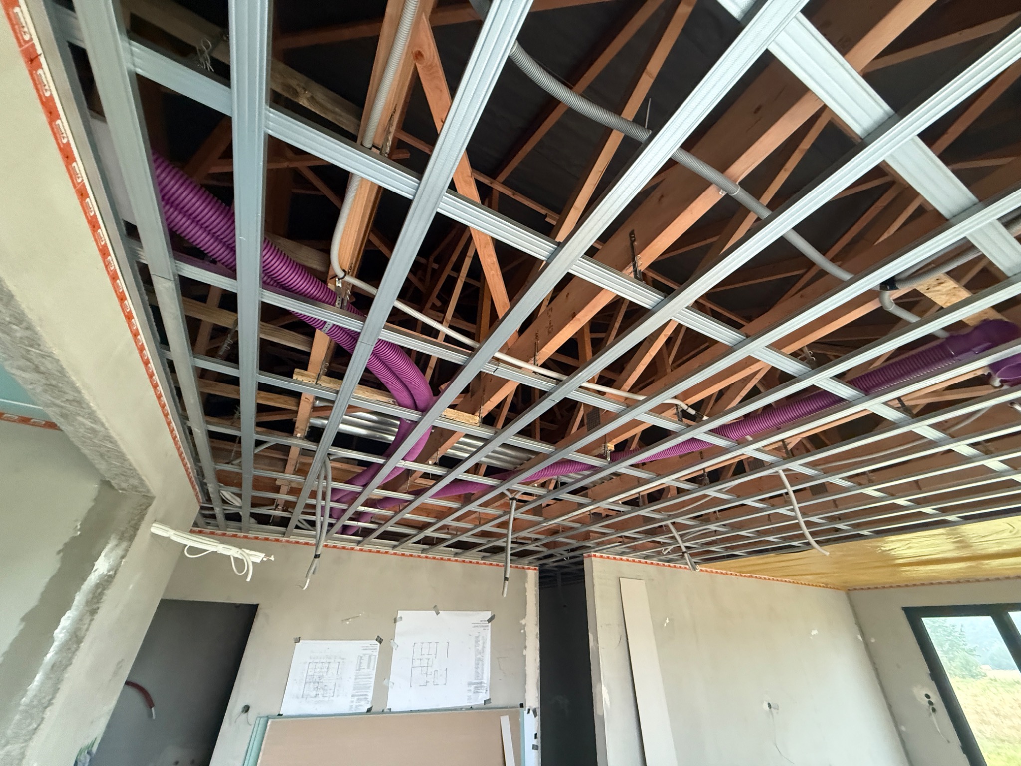 Montaż sufitu podwieszanego z widoczną konstrukcją metalową, rurami wentylacyjnymi i elementami drewnianymi w surowym wnętrzu z planami na ścianie.