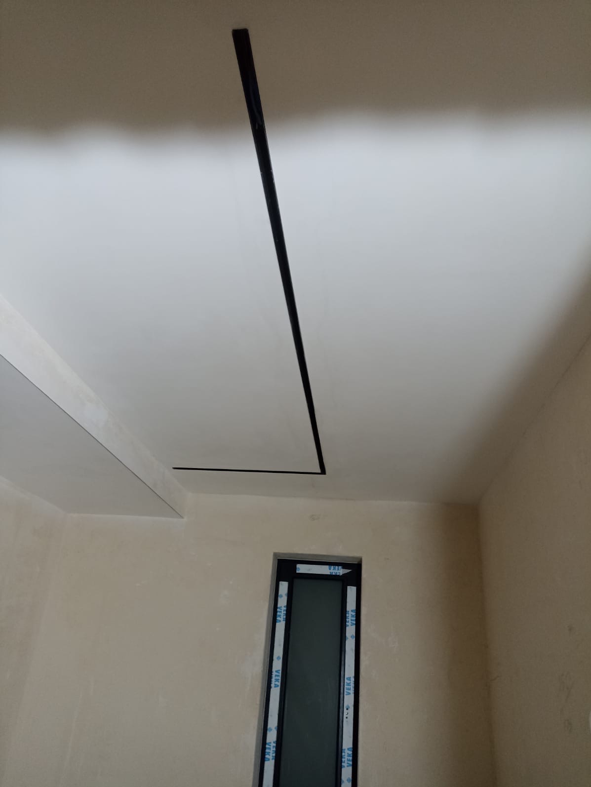 Biały sufit z czarnymi, minimalistycznymi listwami LED, widok z dołu, fragment okna w tle. Neutralne ściany, surowy stan wykończenia.