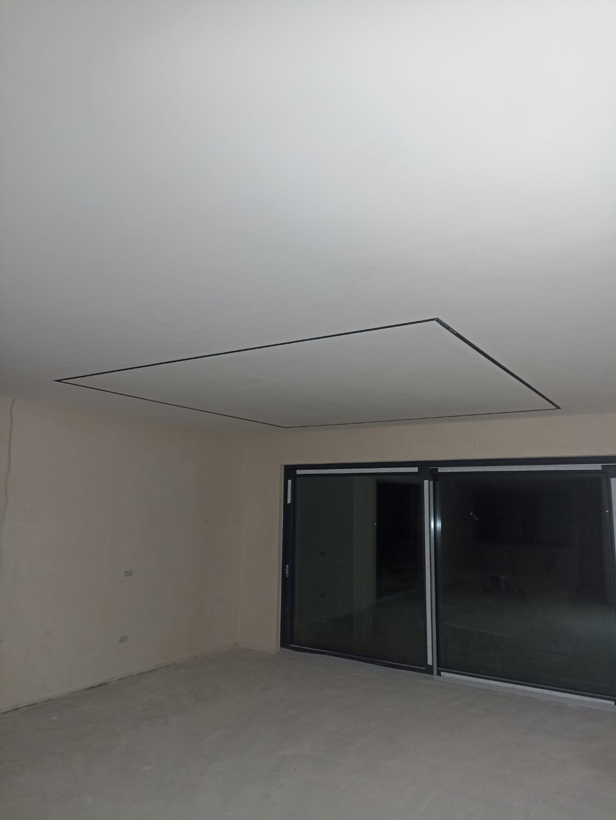 Wykończony sufit z czarną ramką w nowym pomieszczeniu z dużym oknem przesuwnym. Ściany w jasnym odcieniu beżu, podłoga szara. Minimalistyczny design.