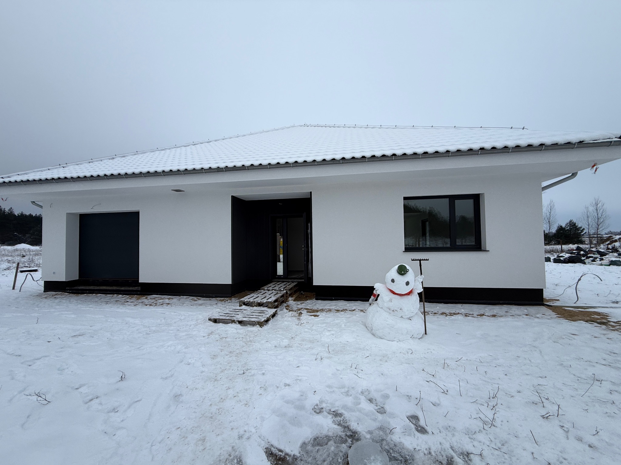 Nowoczesny, parterowy dom z białymi ścianami i ciemnymi oknami w zimowej scenerii. Przed wejściem stoi bałwan z zielonym kubkiem na głowie i grabiami.