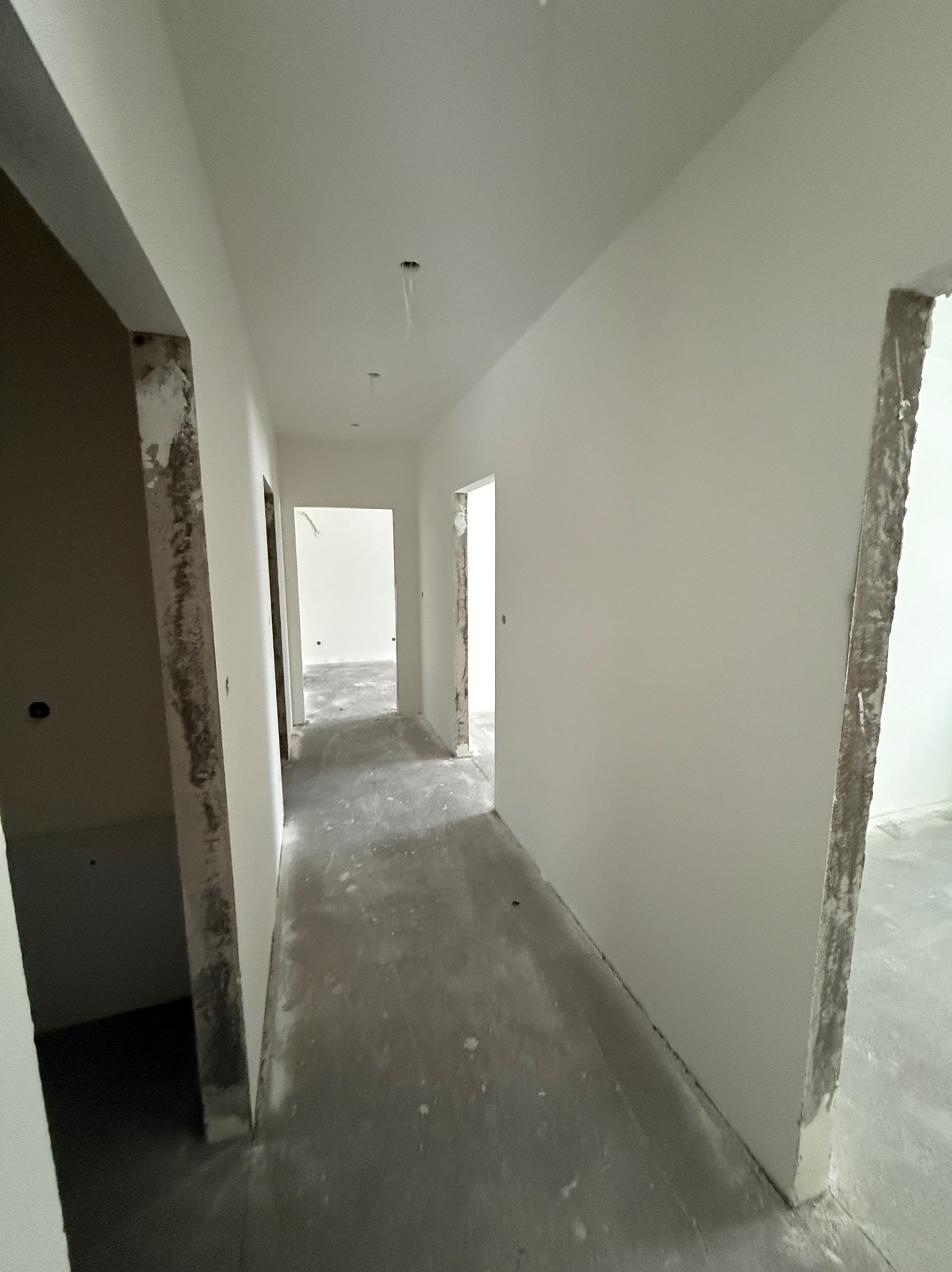 Długi korytarz w trakcie prac wykończeniowych. Ściany świeżo pokryte gładzią, surowy beton na podłodze, widoczne niedokończone ościeżnice. Minimalistyczny, surowy styl.