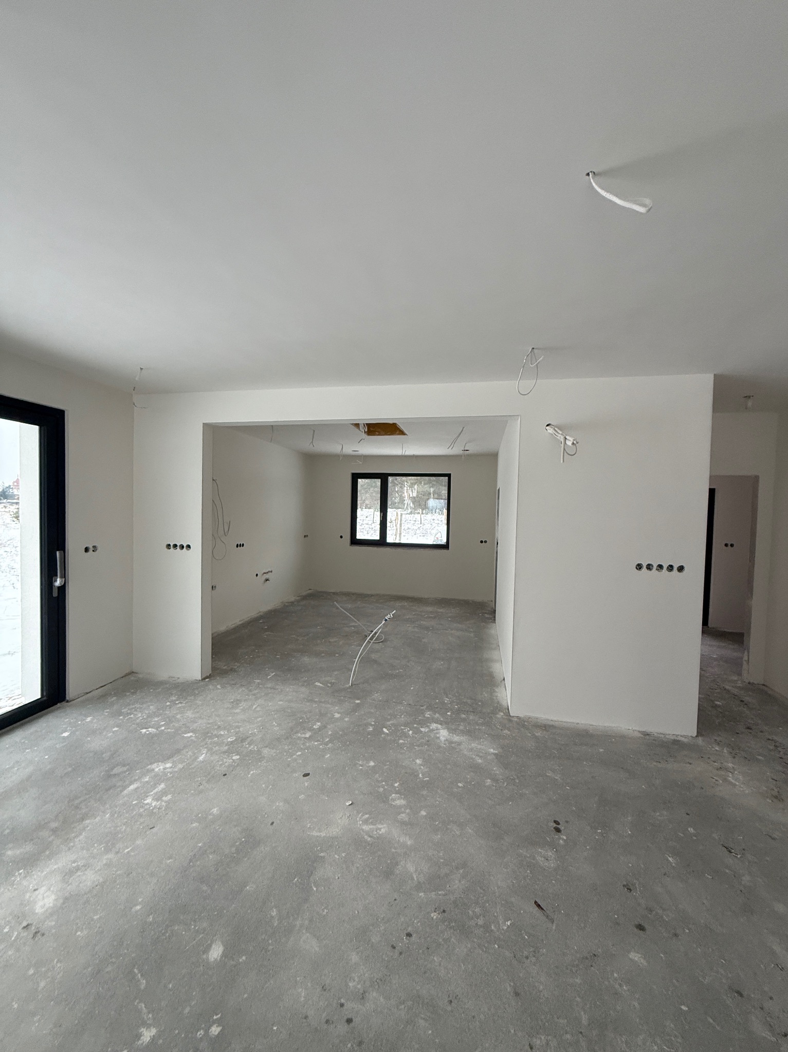Surowe wnętrze domu w trakcie prac wykończeniowych: białe ściany, betonowa podłoga, widoczne kable i puszki elektryczne. Okno z czarną ramą w tle.