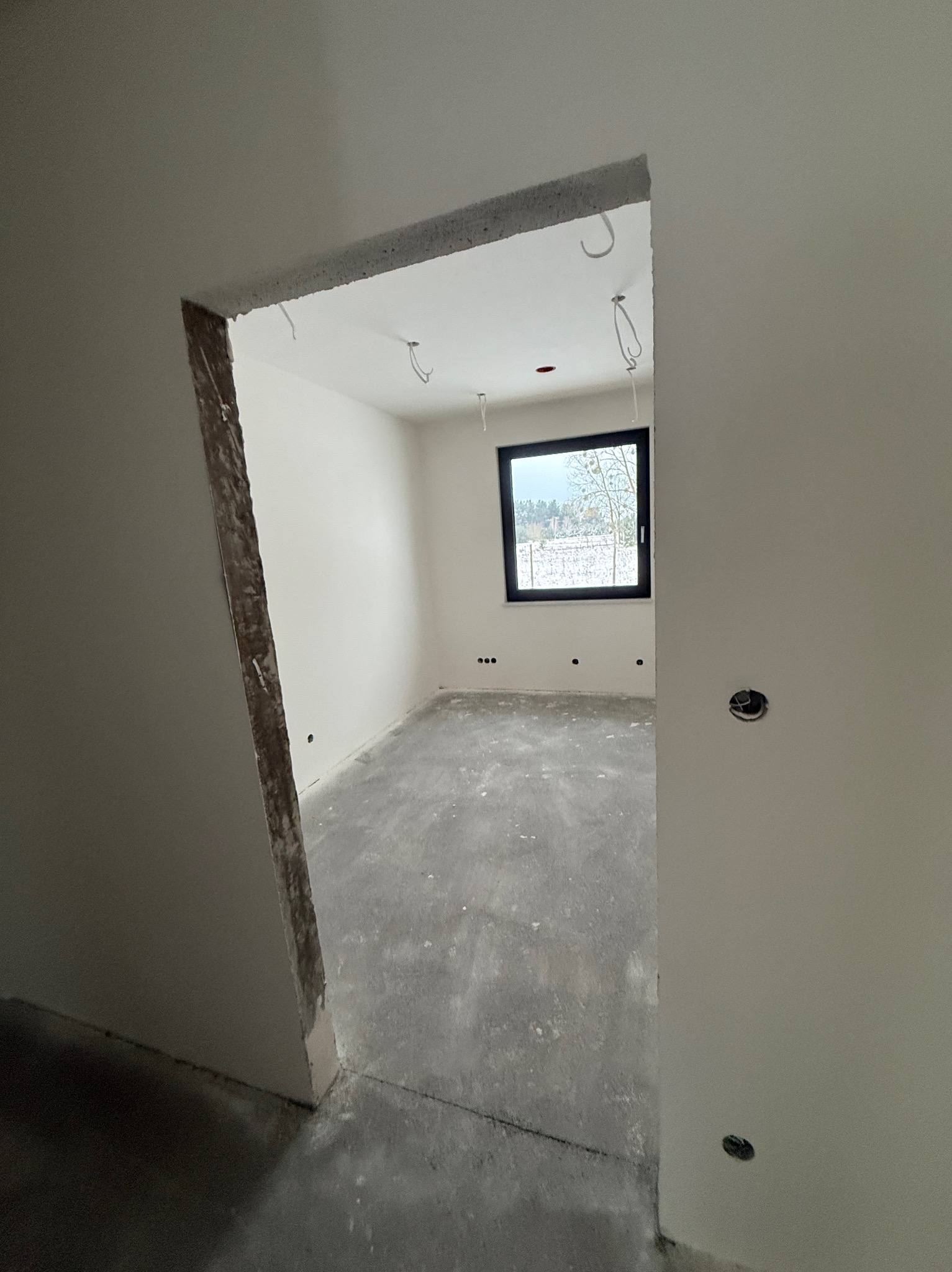 Surowe wnętrze z oknem, widoczne kable i niedokończone ściany. W tle zimowy krajobraz za oknem. Wnętrze w trakcie prac wykończeniowych, widok z perspektywy wejścia.