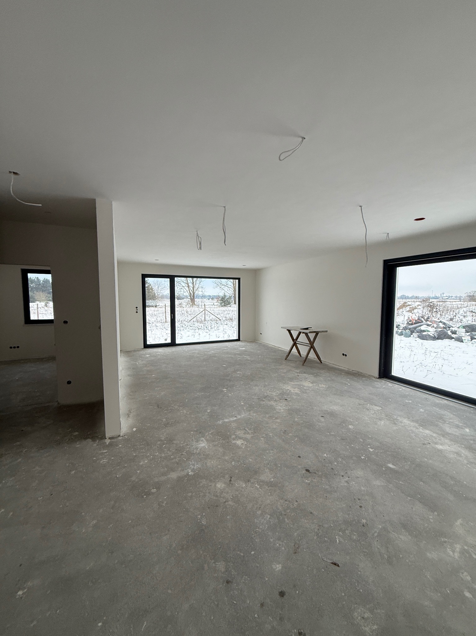 Surowe wnętrze z dużymi oknami i betonową podłogą, przygotowane do dalszych prac wykończeniowych. Widoczne wystające kable elektryczne i prowizoryczny stolik.
