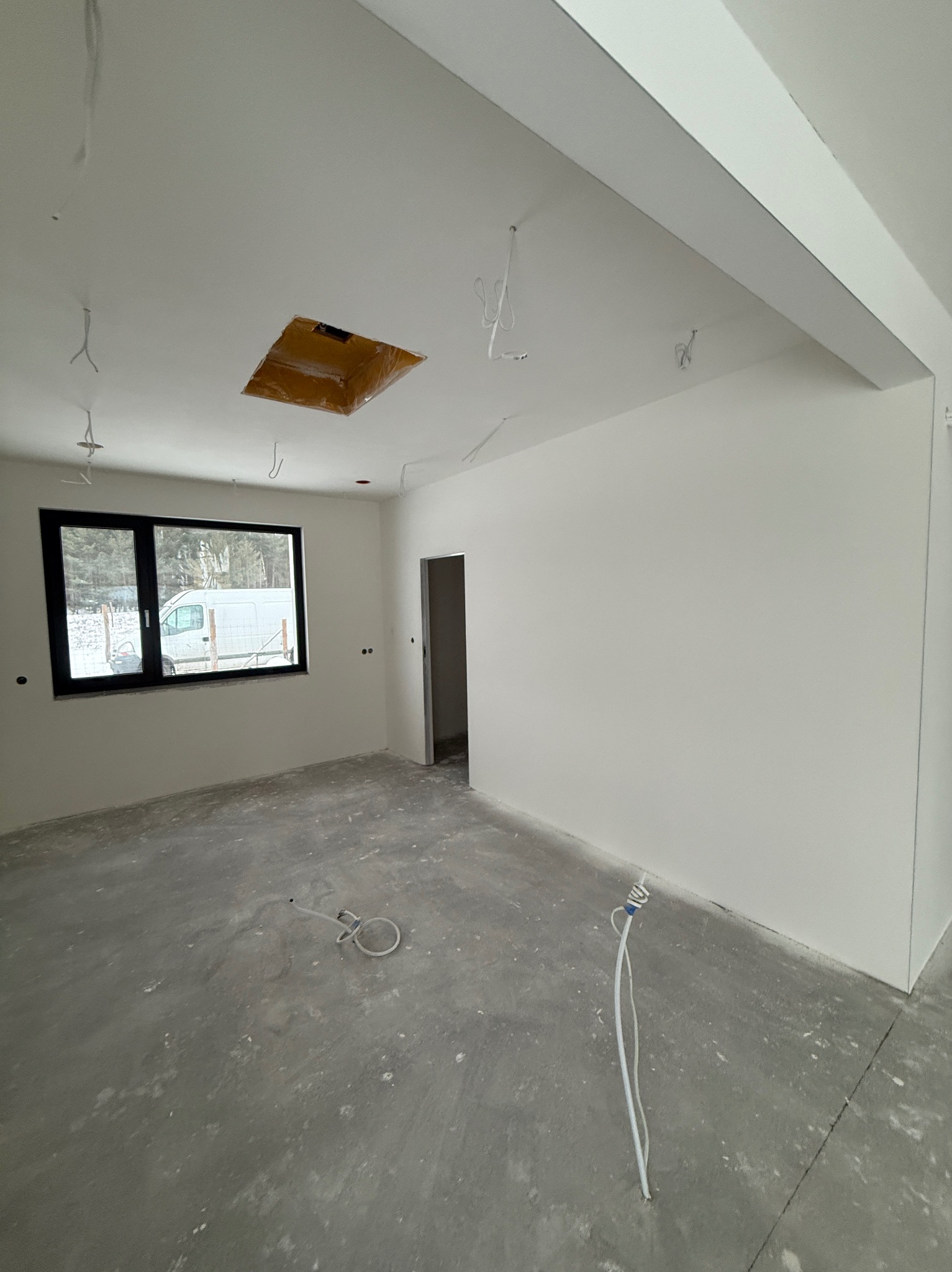 Wnętrze w trakcie wykończenia: białe ściany, surowa podłoga, okno z widokiem na zewnątrz, wystające kable elektryczne. Otwór w suficie zabezpieczony folią.