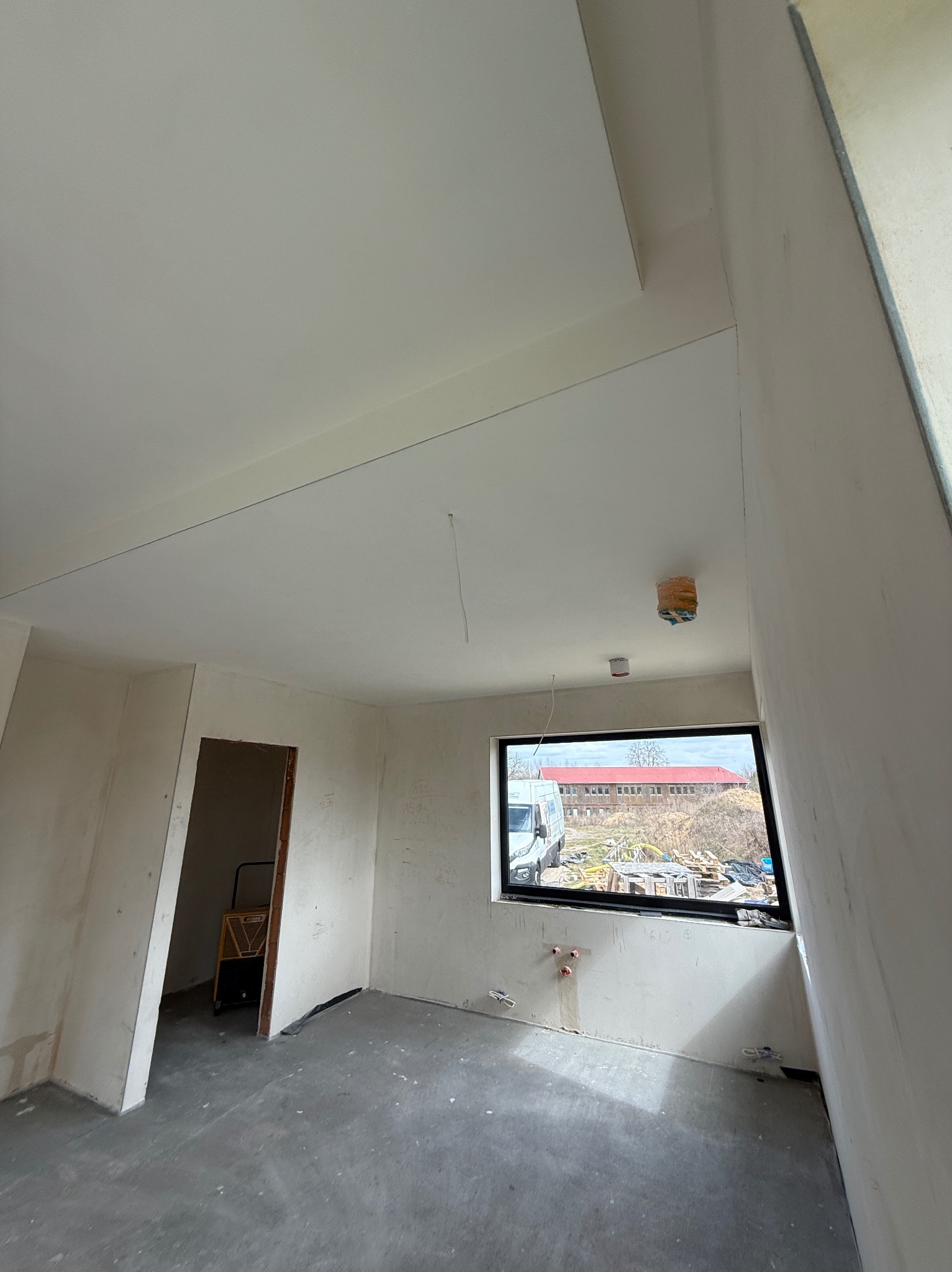 Wnętrze w trakcie prac wykończeniowych: surowe ściany, gładki sufit z widocznymi przewodami, okno z widokiem na zewnątrz, betonowa podłoga. W tle drabina.