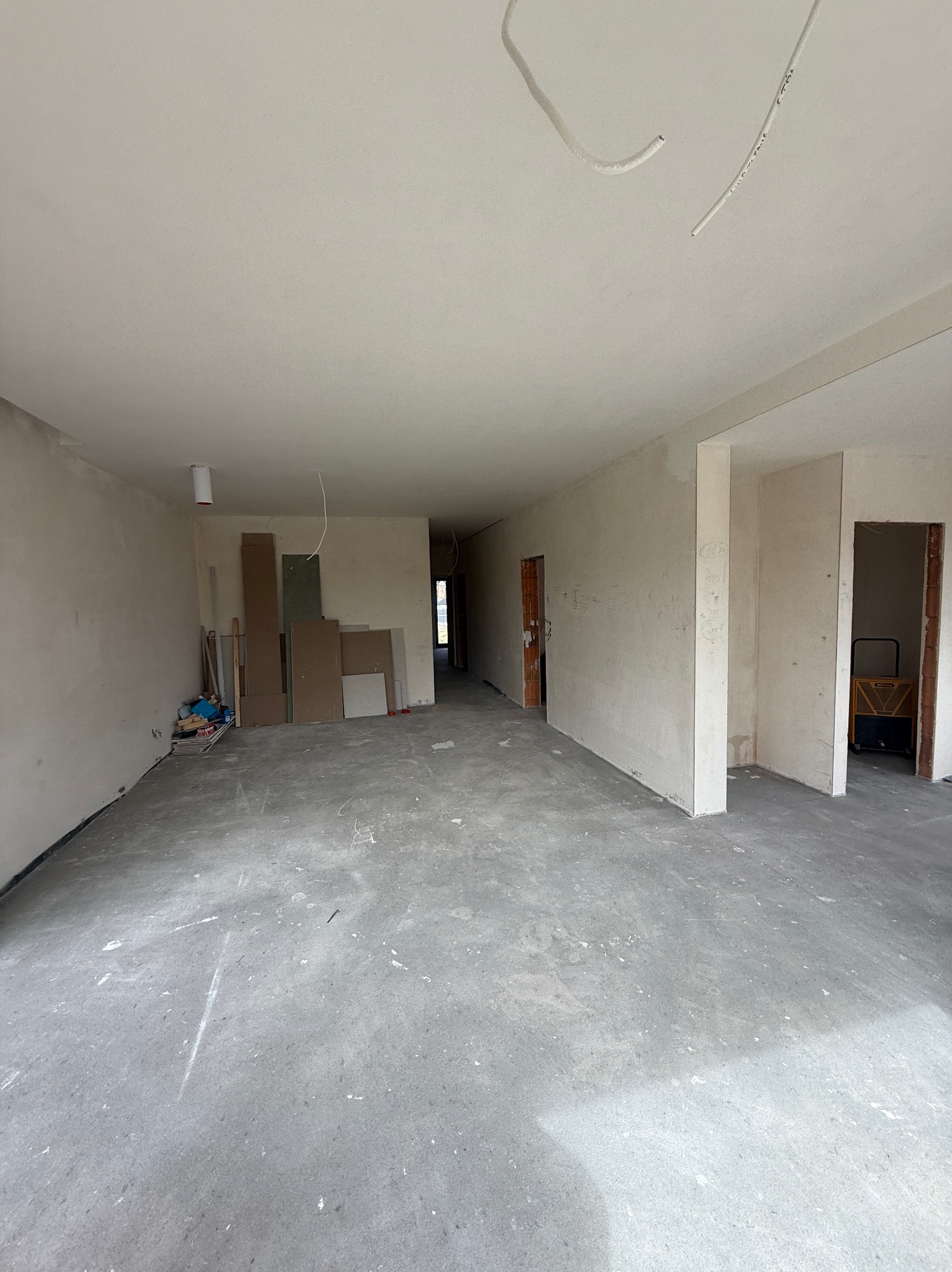 Surowe wnętrze w trakcie prac: szare ściany, betonowa podłoga, widoczne kable i materiały budowlane. Przestrzeń przygotowana do dalszych prac wykończeniowych.
