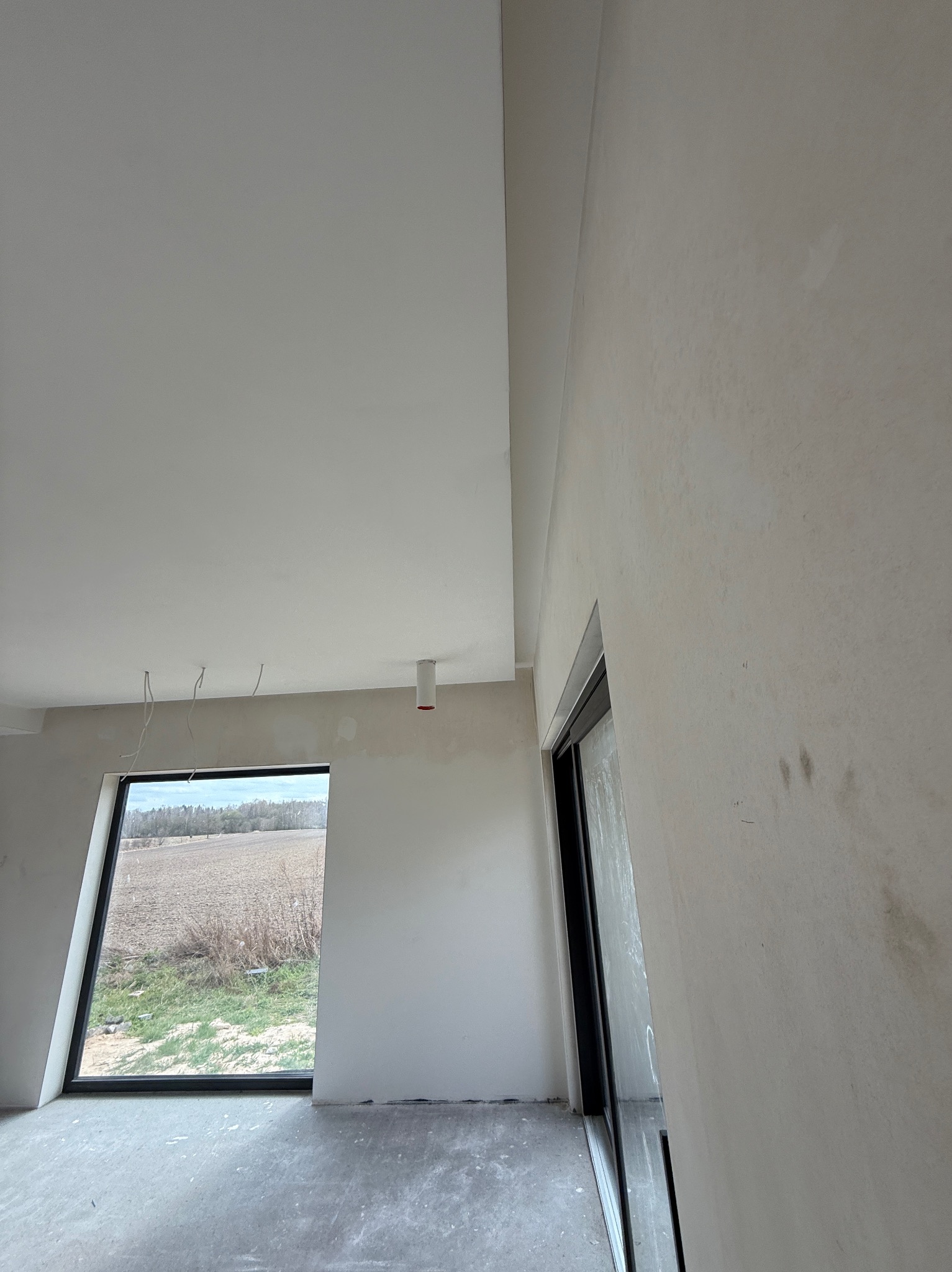 Wnętrze w trakcie prac wykończeniowych: ściany przygotowane pod malowanie, widoczne przewody elektryczne, okno z widokiem na pole. Minimalistyczny styl.