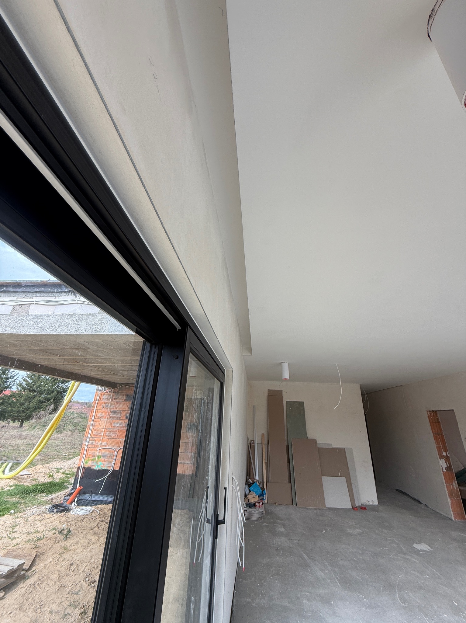 Wnętrze w trakcie prac wykończeniowych: gładkie ściany i sufit, widoczne płyty karton-gips, okno z czarną ramą. Na zewnątrz widoczny teren budowy.