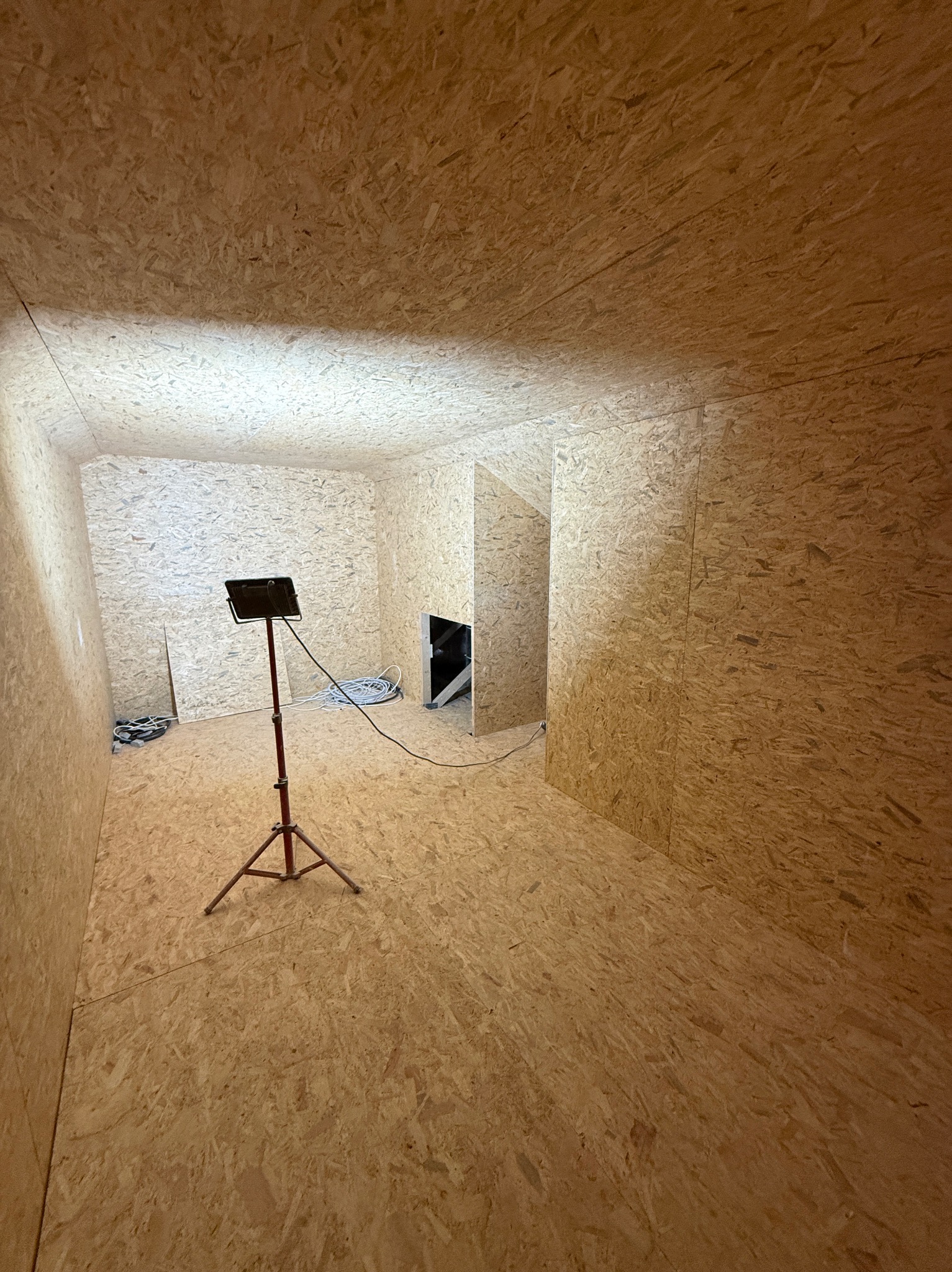 Pokój wyłożony płytami OSB, oświetlony lampą na statywie. Widoczne kable i fragmenty konstrukcji. Przygotowanie do dalszych prac wykończeniowych.