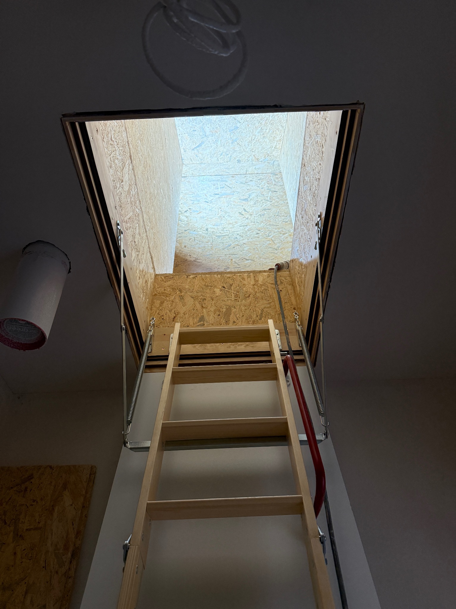 Otwór strychowy z drewnianą składaną drabinką, widok z dołu. Widoczne płyty OSB i sprężyny mechanizmu. Ujęcie z perspektywy żabiej.