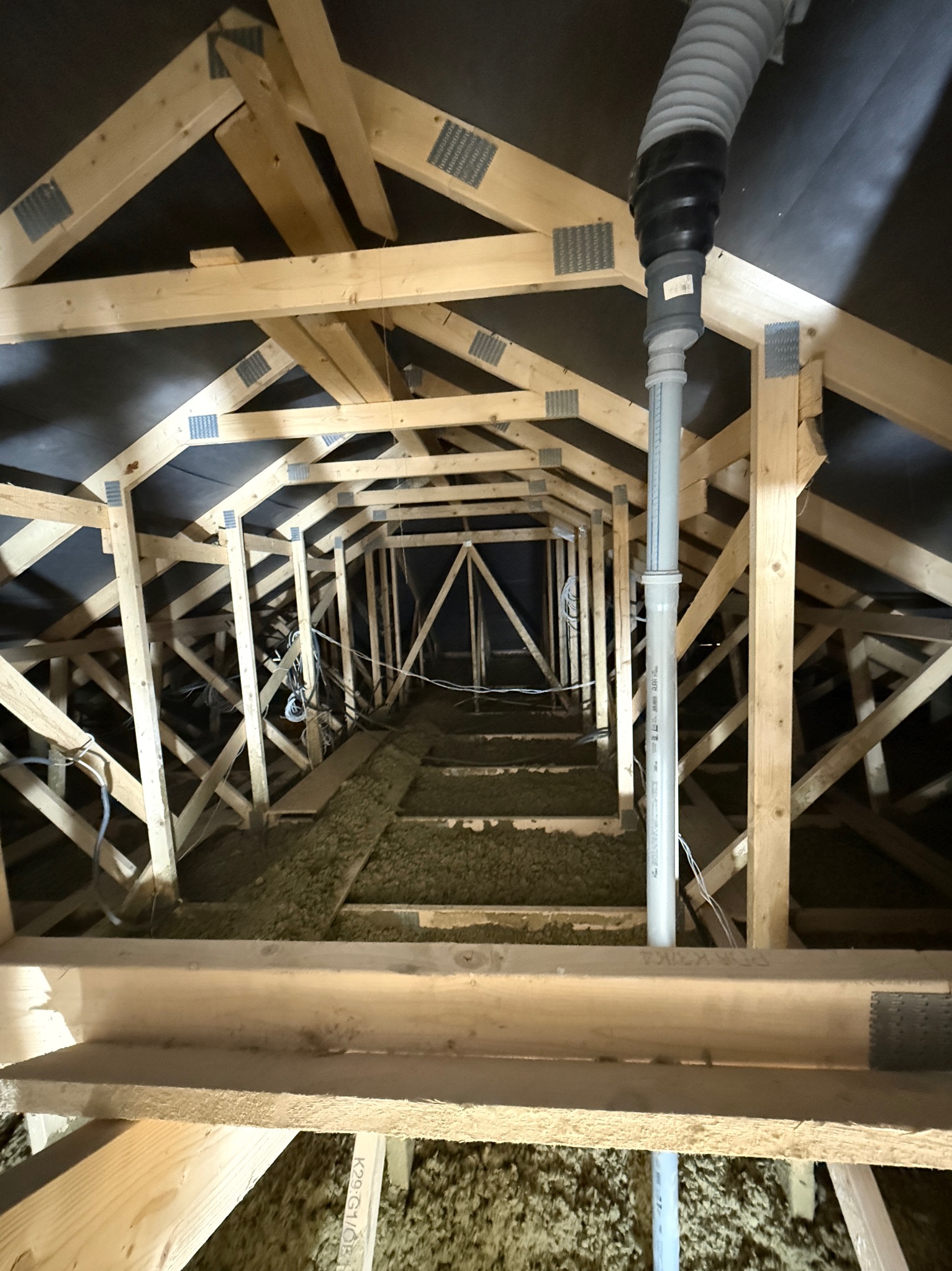 Szkielet dachu z izolacją z wełny mineralnej, widok z dołu. Elementy drewniane łączone metalowymi płytkami. Rura wentylacyjna pionowo w kadrze.