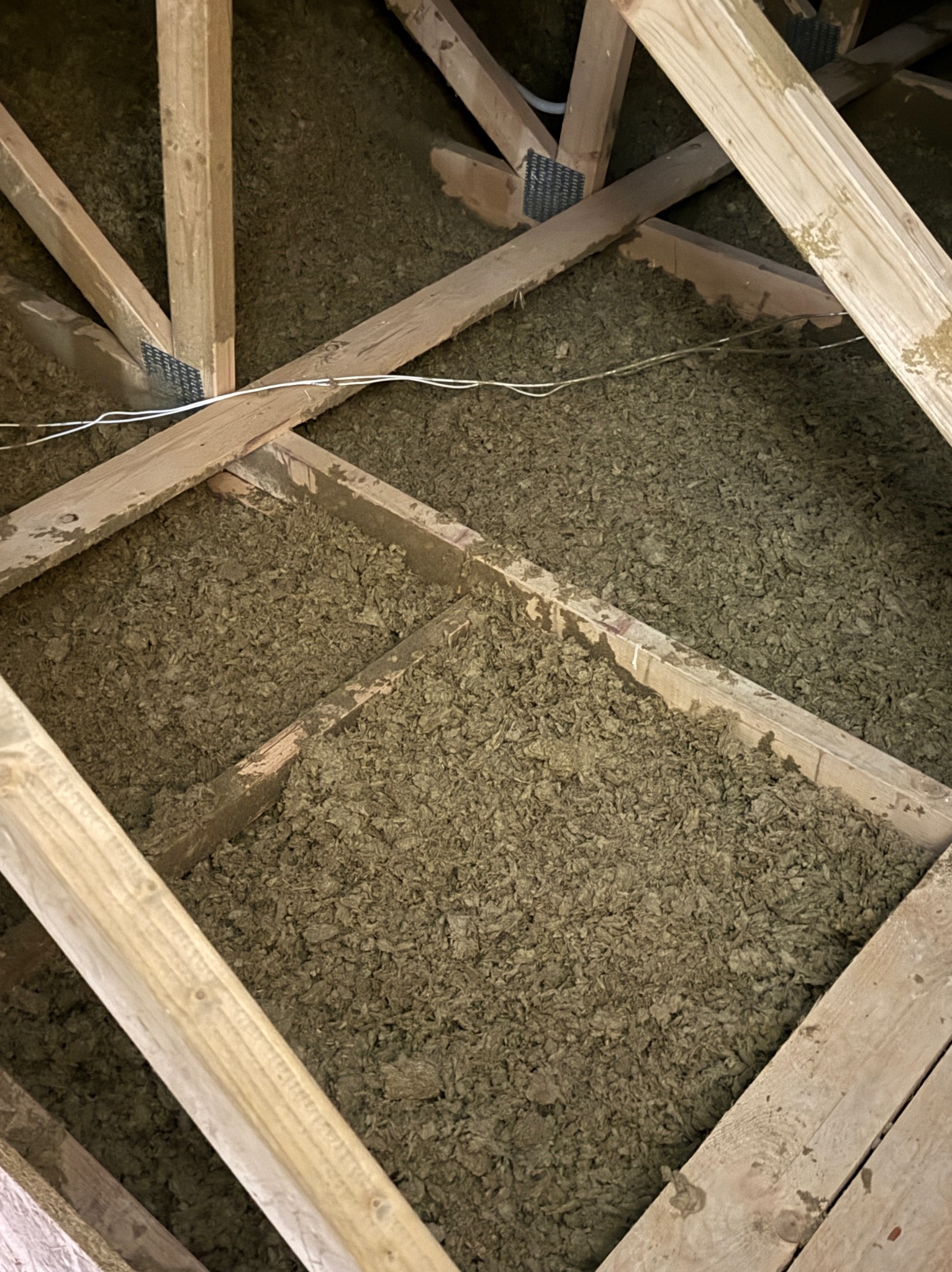 Ocieplenie poddasza wełną mineralną, widoczne drewniane elementy konstrukcyjne i biały przewód elektryczny. Ujęcie z góry, wypełnienie przestrzeni izolacją.