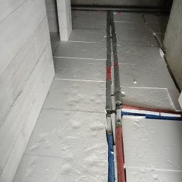 Dawid Koziołek - Izolacja fundamentów z białego styropianu, widoczne rury instalacyjne w kolorze niebieskim i czerwonym, surowy betonowy strop.