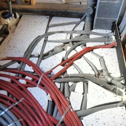 Dawid Koziołek - Przygotowanie podłoża pod wylewkę: fragment podłogi z ułożonymi instalacjami grzewczymi w czerwonych rurach, szarymi rurami kanalizacyjnymi i fragmentami białego styropianu izolacyjnego między nimi...