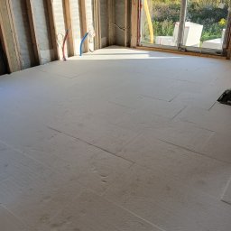 Dawid Koziołek - Wnętrze w stanie surowym z izolacją podłogową z płyt styropianowych, widoczne odsłonięte drewniane elementy konstrukcyjne ścian, rurki instalacji oraz okno z widokiem na krajobraz.