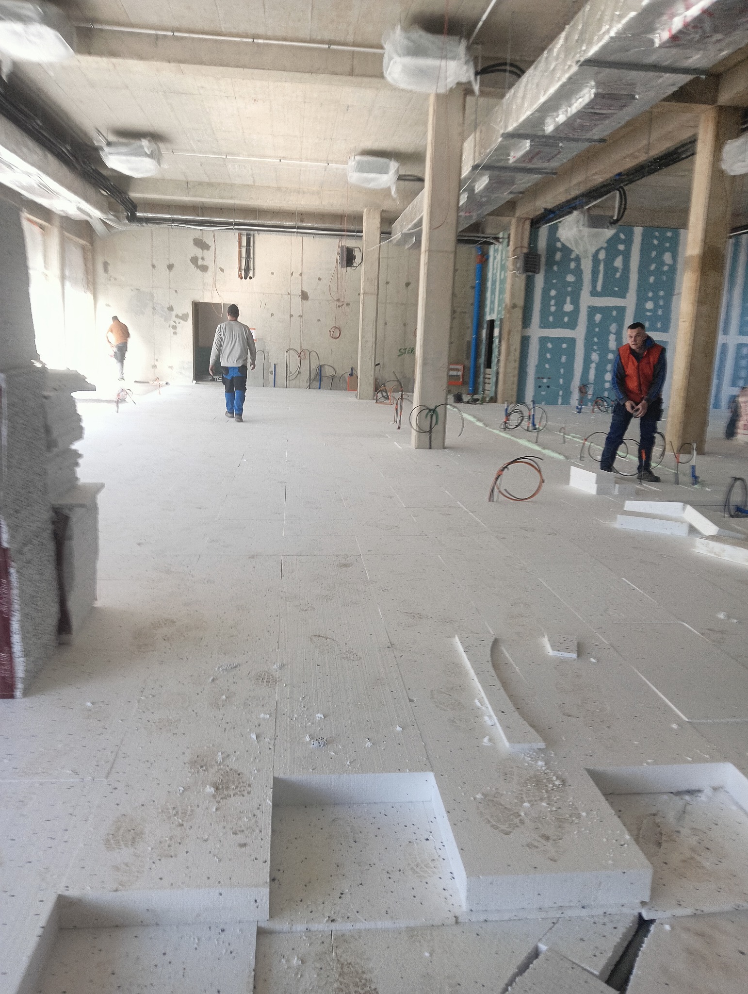 Wnętrze w trakcie prac budowlanych: układanie białych płyt izolacyjnych na podłodze, widoczne kable instalacji, betonowe kolumny i pracownicy.
