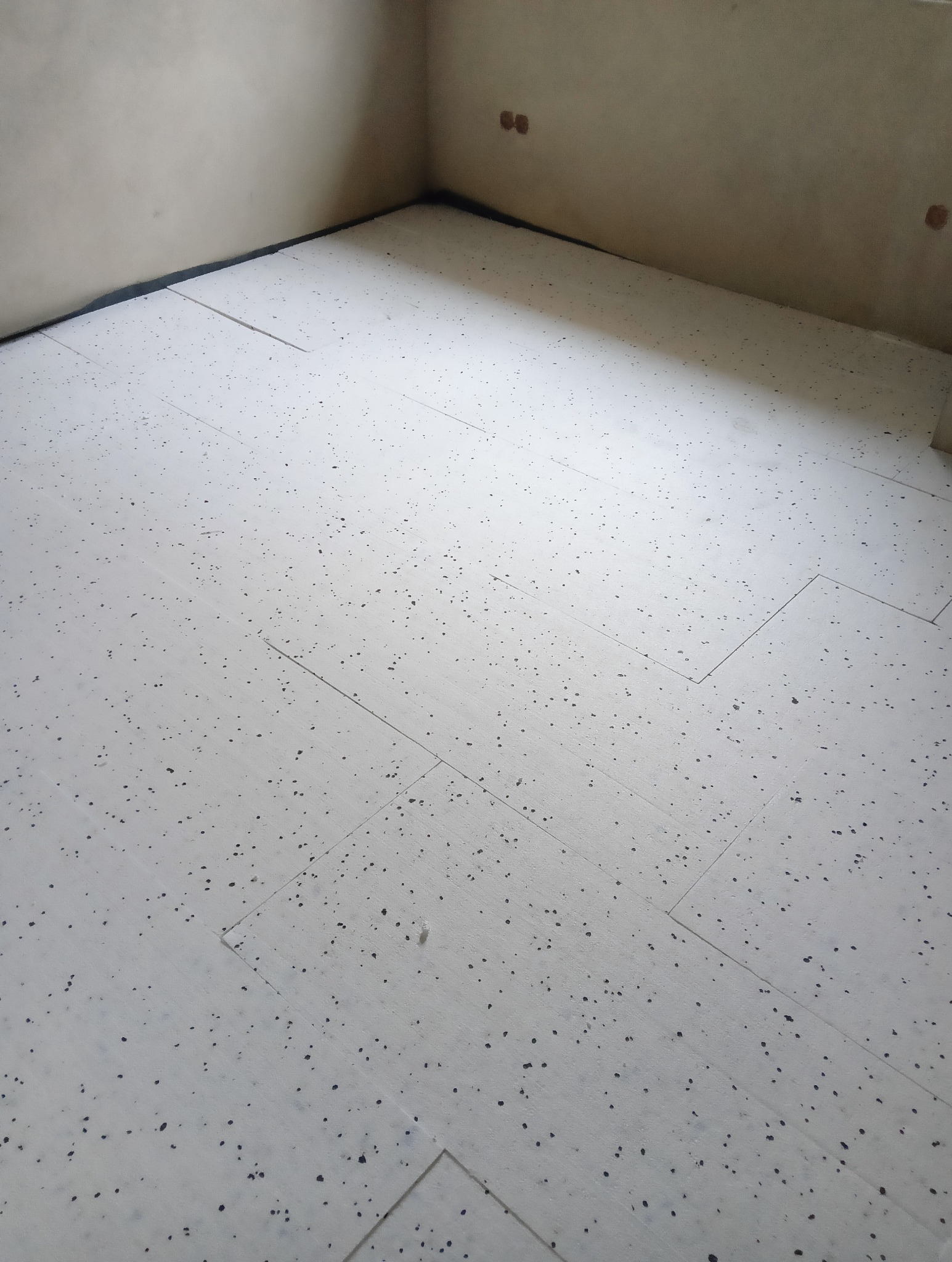 Białe płyty izolacyjne z czarnymi plamkami ułożone na podłodze w rogu pomieszczenia, widoczne ślady po gniazdkach elektrycznych na ścianie.