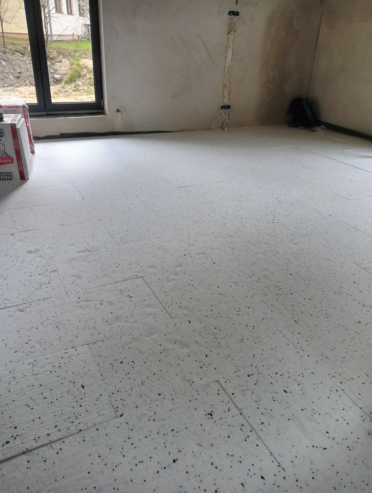 Wnętrze w trakcie prac wykończeniowych, widoczna podłoga pokryta płytami styropianowymi z czarnymi granulkami, przygotowana pod wylewkę, ściany surowe, widoczne puszki elektryczne, okno w tle.