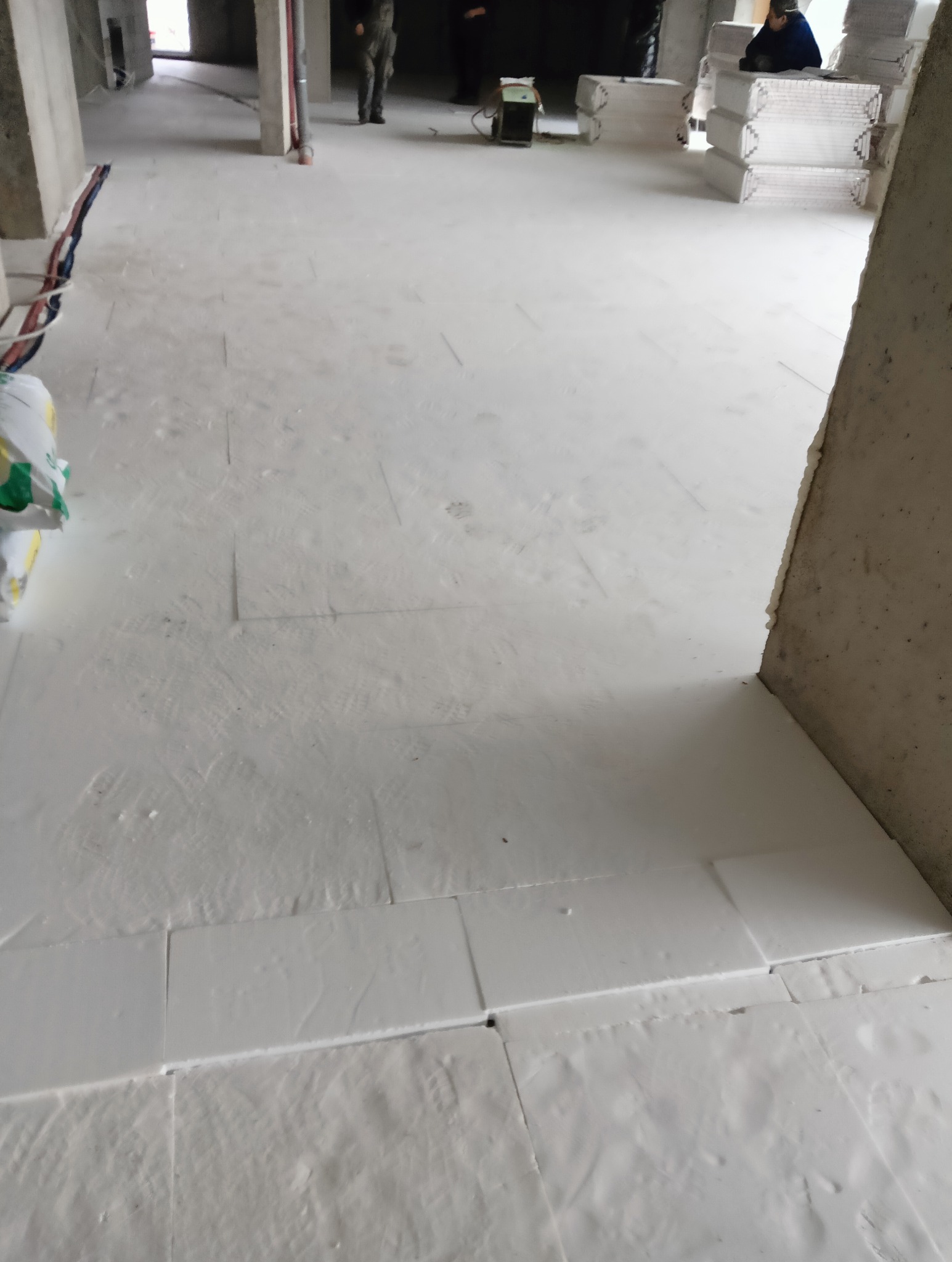 Wnętrze w trakcie prac wykończeniowych: biała, gładka posadzka z widocznymi łączeniami płyt, worki z materiałami budowlanymi w rogu, osoby w tle i sprzęt budowlany.