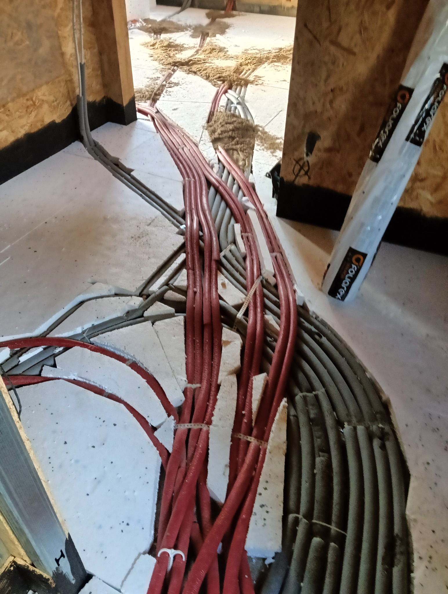 Rury grzewcze podłogowe, czerwone i szare, układane w warstwie styropianu z fragmentami jastrychu, widoczne elementy izolacji pionowej przy ścianie i opakowanie produktu marki Fouarex.