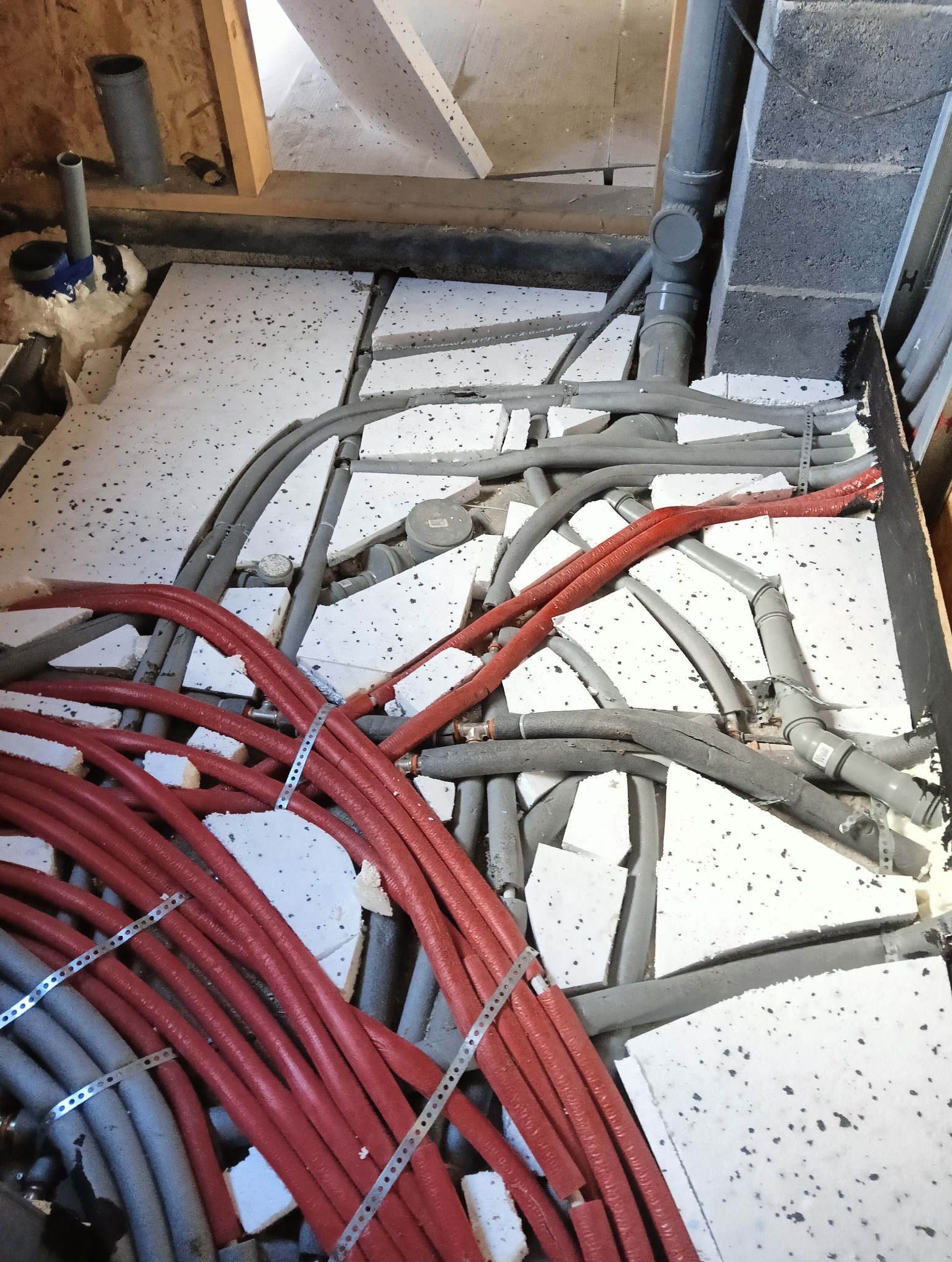 Przygotowanie podłoża pod wylewkę: fragment podłogi z ułożonymi instalacjami grzewczymi w czerwonych rurach, szarymi rurami kanalizacyjnymi i fragmentami białego styropianu izolacyjnego między nimi...