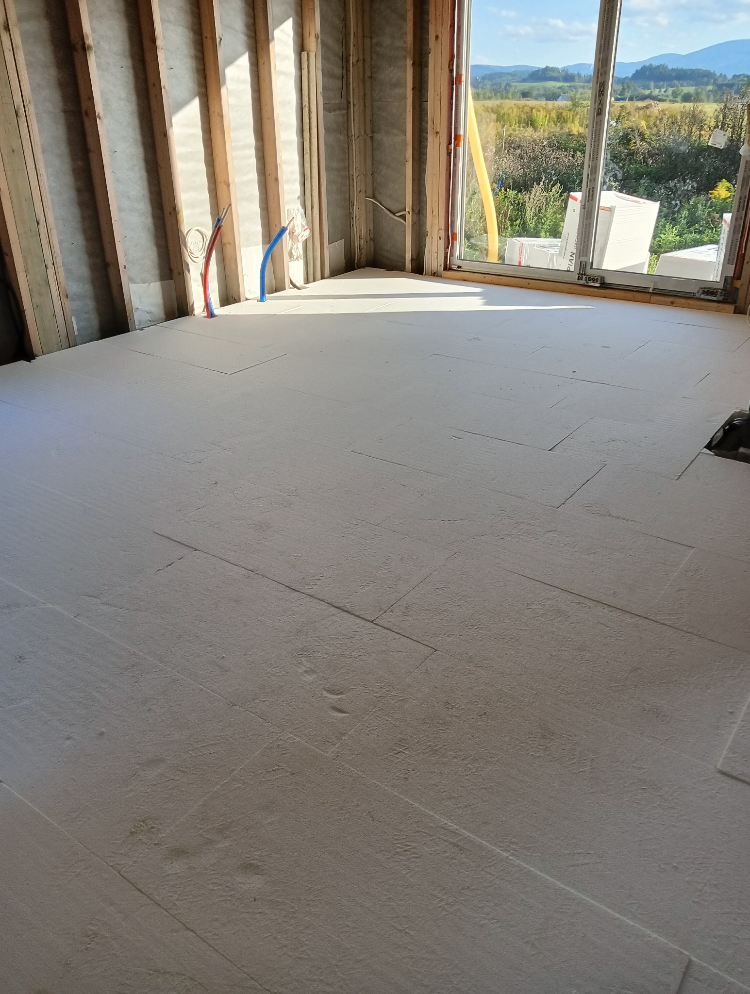 Wnętrze w stanie surowym z izolacją podłogową z płyt styropianowych, widoczne odsłonięte drewniane elementy konstrukcyjne ścian, rurki instalacji oraz okno z widokiem na krajobraz.