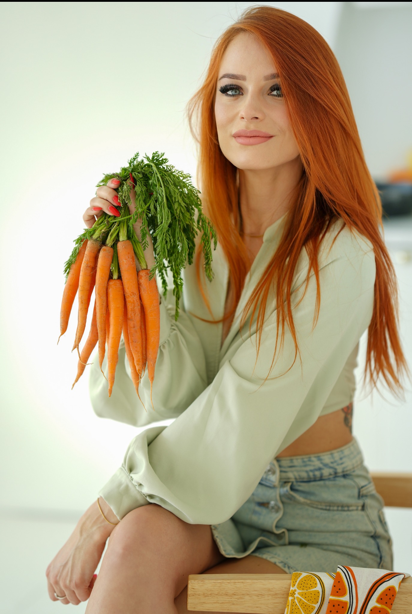 Uśmiechnięta kobieta z rudymi włosami trzyma pęczek świeżych marchwi z zielonymi liśćmi na jasnym tle. Zdjęcie promuje zdrowe odżywianie i świeże warzywa.