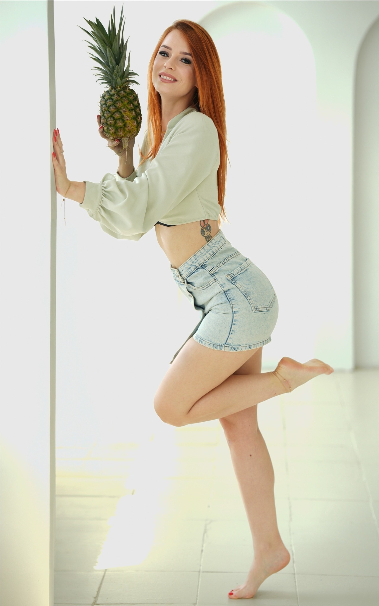 Uśmiechnięta, ruda kobieta opiera się o białą ścianę, trzymając ananasa. Ma na sobie dżinsowe szorty i bluzkę odsłaniającą brzuch. Widoczny tatuaż.