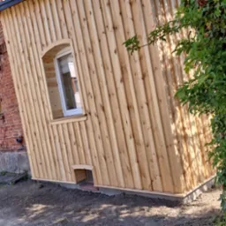 Nowa drewniana elewacja budynku z oknem w kształcie łuku, przylegająca do starego budynku z czerwonej cegły, widoczna zarośnięta roślinność.