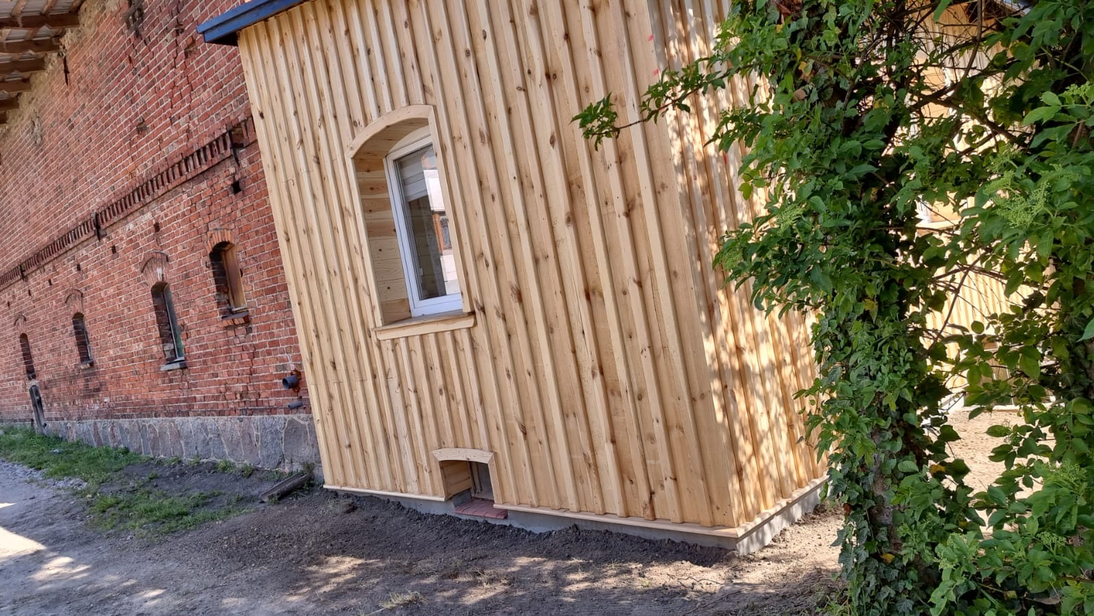 Nowa drewniana elewacja budynku z oknem w kształcie łuku, przylegająca do starego budynku z czerwonej cegły, widoczna zarośnięta roślinność.