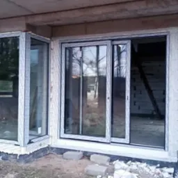 Narożne okno i przesuwne okno tarasowe z białymi ramami PCV w surowym stanie budynku z białego bloczka.