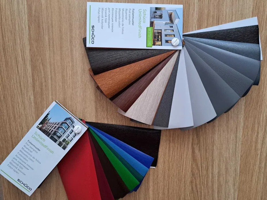 Dwa wachlarze próbek oklein marki Schüco w różnych kolorach i fakturach, ułożone na drewnianym blacie, prezentujące bogatą paletę wykończeń.