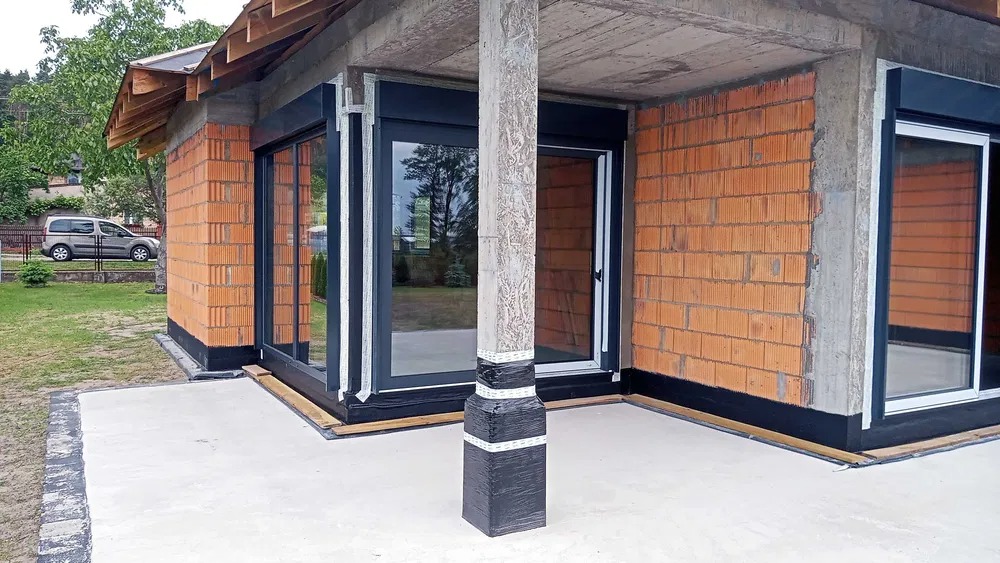 Surowy budynek w trakcie budowy z zamontowanymi oknami i roletami zewnętrznymi w czarnych ramach, widoczne cegły i beton, kolumna zabezpieczona folią.