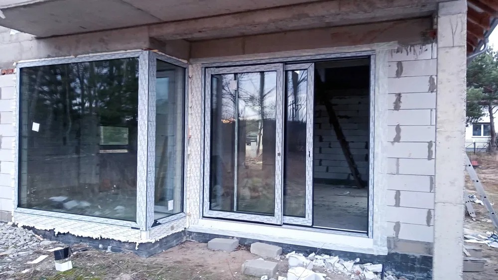 Narożne okno i przesuwne okno tarasowe z białymi ramami PCV w surowym stanie budynku z białego bloczka.