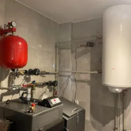 Kotłownia w jasnych płytkach: kocioł grzewczy, czerwony zbiornik wyrównawczy, biały bojler oraz rury instalacji centralnego ogrzewania z zaworami i pompami obiegowymi.