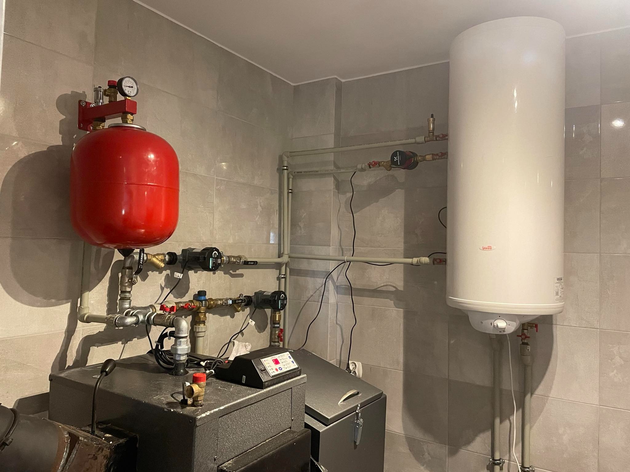 Kotłownia w jasnych płytkach: kocioł grzewczy, czerwony zbiornik wyrównawczy, biały bojler oraz rury instalacji centralnego ogrzewania z zaworami i pompami obiegowymi.