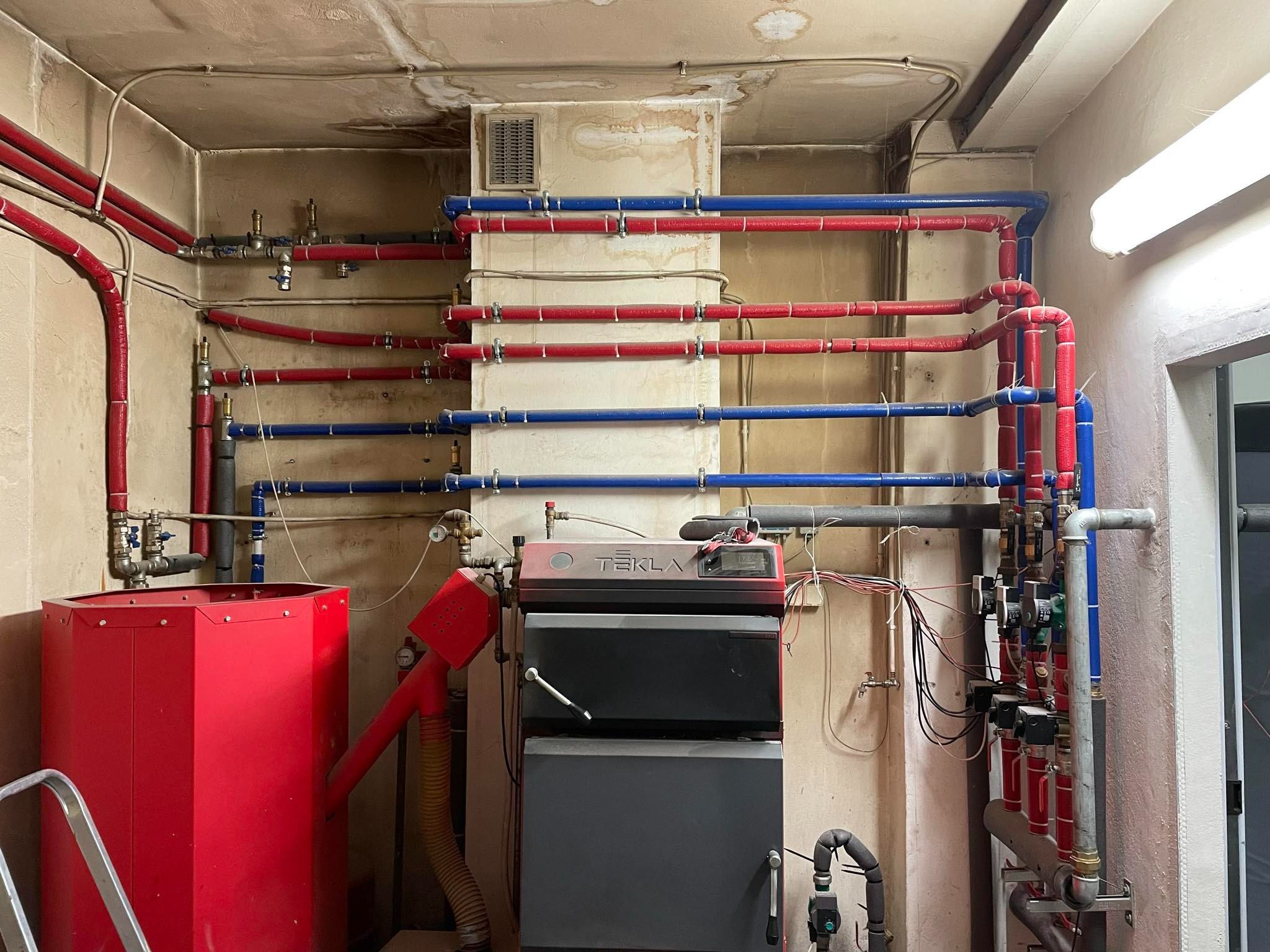 Kotłownia z piecem Tekla, czerwonym zasobnikiem i rozbudowaną instalacją rur w kolorach czerwonym i niebieskim, widoczne ślady wilgoci na suficie i ścianach.