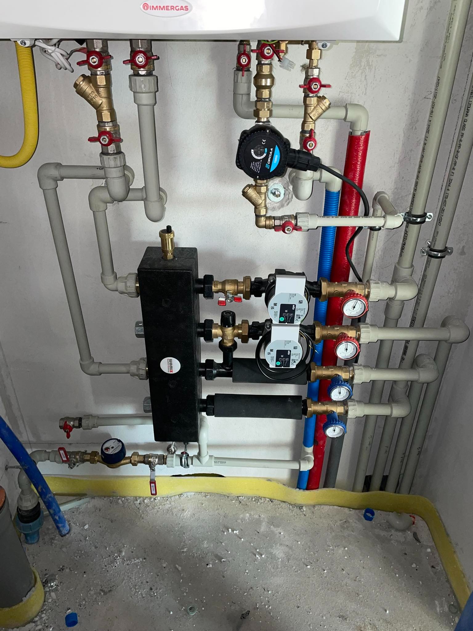 Kompleksowa instalacja hydrauliczna z piecem gazowym Immergas, widoczne pompy obiegowe Wilo, czarny rozdzielacz Womix, rury PEX w kolorach szarym, czerwonym i niebieskim, manometry z czerwonymi...