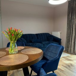 Volodymyr Dubov INVO-Bud - Nowoczesny salon z okrągłym drewnianym stołem, niebieskimi krzesłami z weluru i sofą w tle, oświetlony okrągłą lampą sufitową, z bukietem czerwonych tulipanów w wazonie na stole.