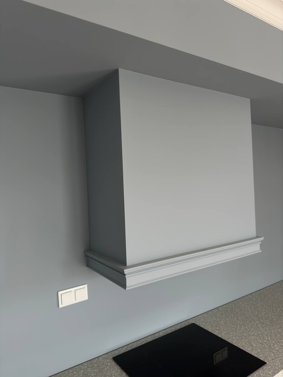 Szary, zabudowany okap kuchenny z ozdobną listwą, dopasowany kolorystycznie do ściany, nad płytą indukcyjną; widoczny włącznik światła na ścianie.