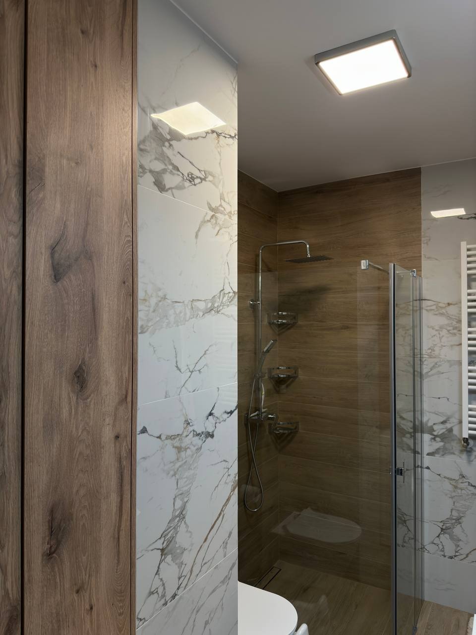 Nowoczesna łazienka z kabiną prysznicową typu walk-in, wykończona płytkami imitującymi marmur Carrara i drewno, oświetlona kwadratowymi lampami sufitowymi.