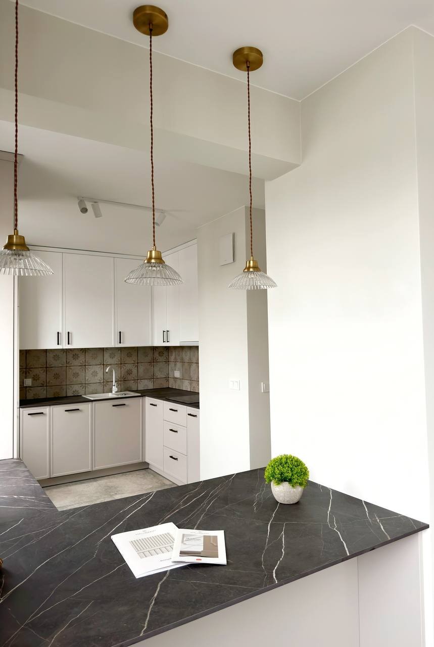 Nowoczesna, jasna kuchnia w Warszawie z białymi szafkami, czarnym blatem z marmurowym wzorem, mozaiką nad blatem roboczym i trzema wiszącymi lampami z kloszami z żłobionego szkła.