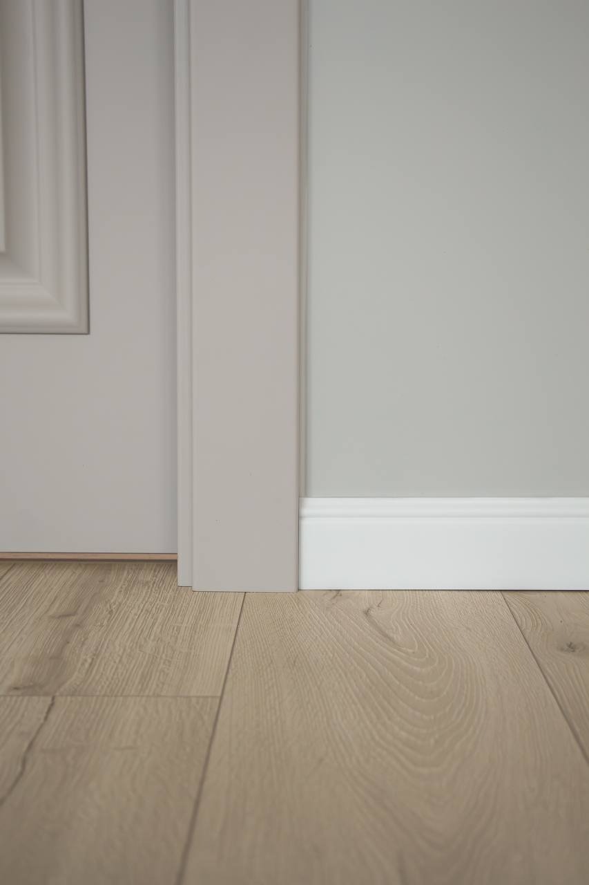 Fragment wnętrza z jasną drewnianą podłogą, białą listwą przypodłogową i szarą ścianą, widoczny fragment ościeżnicy drzwi.