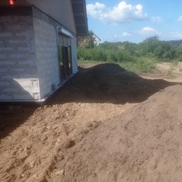 Bartosz Lenkiewicz usługi budowlano elektryczne - Surowy stan budynku z betonu komórkowego z widoczną izolacją fundamentów i pryzmą ziemi wokół, na tle zieleni i błękitnego nieba.