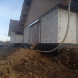 Bartosz Lenkiewicz usługi budowlano elektryczne - Budynek w trakcie ocieplania, widoczne ściany z bloczków, izolacja fundamentów z folii kubełkowej oraz rura do wdmuchiwania izolacji, sterta ziemi na pierwszym planie.