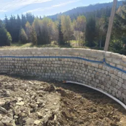 Niski, zakrzywiony murek z kamienia polnego, z niebieskim kablem elektrycznym przymocowanym na zewnątrz i białą rurą drenażową wewnątrz, na tle ziemi, drzew i gór.