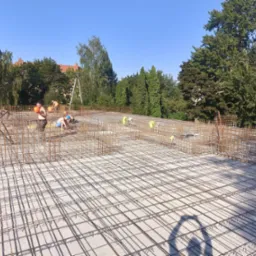 Zbrojenie płyty fundamentowej z widocznymi pracownikami w kaskach podczas montażu siatki zbrojeniowej, w tle drzewa i budynki.