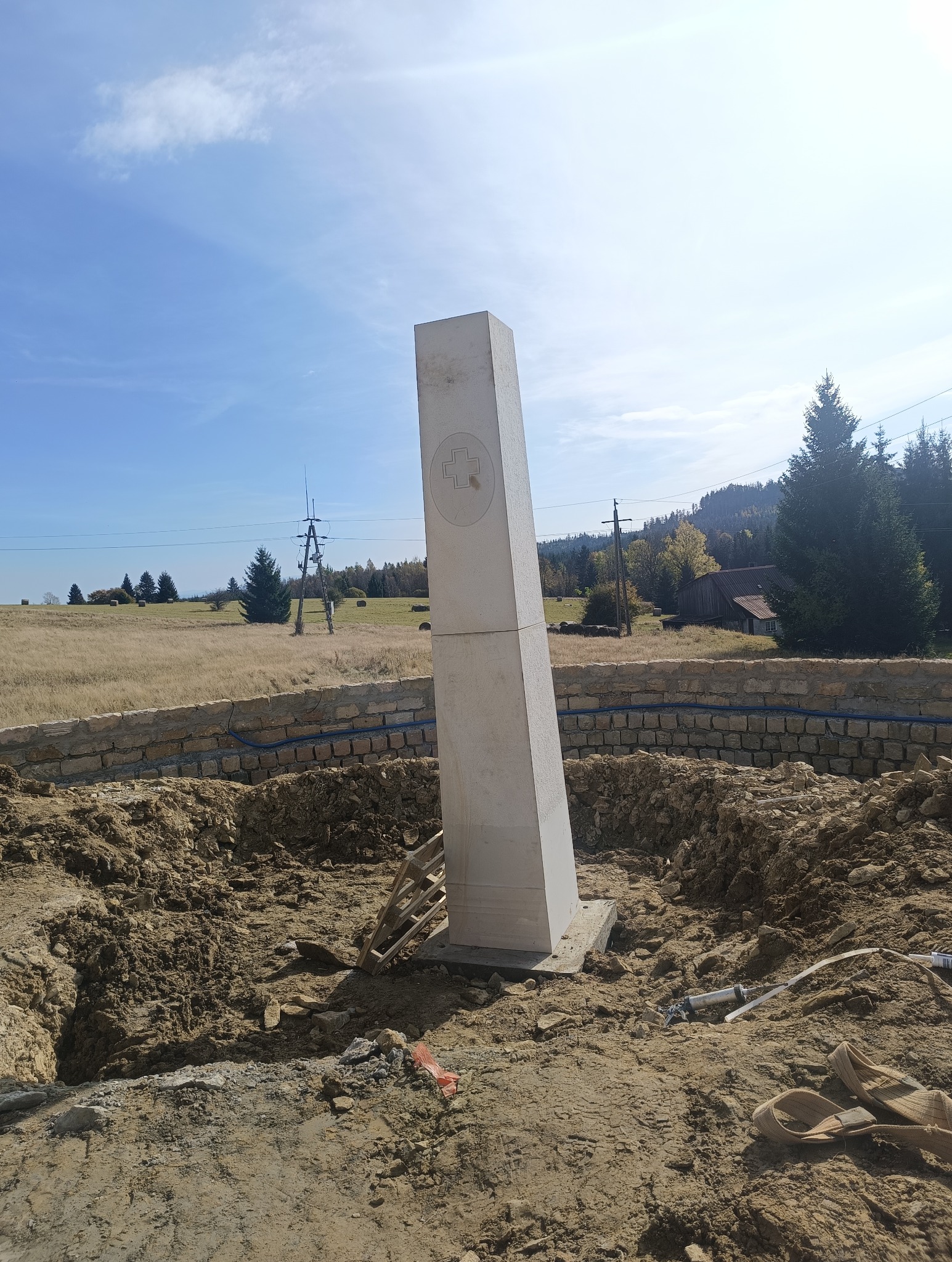 Wzniesiony biały obelisk z wyrytym krzyżem w okręgu, otoczony pierścieniem z kamienia, w trakcie prac ziemnych na tle krajobrazu z polami i lasem.