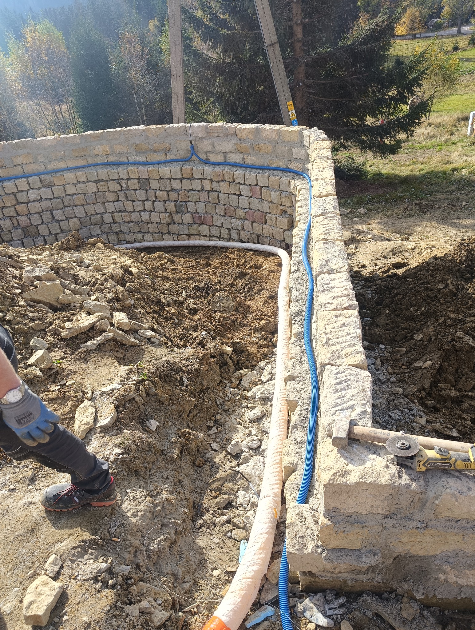Budowa okrągłego fundamentu z kamienia, z widocznymi rurami instalacyjnymi w ziemi i narzędziami murarskimi na murze, w tle drzewa i słup energetyczny.